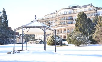 a snow - covered park with a large hotel in the background , surrounded by trees and benches at Falkenstein Grand, Autograph Collection