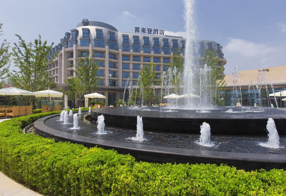 a large building with a fountain in front of it , surrounded by green grass and trees at Sheraton Qinhuangdao Beidaihe  Hotel