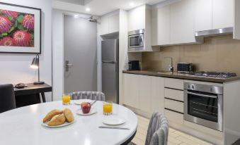 a kitchen with white cabinets , a dining table with chairs , and a television mounted on the wall at Meriton Suites North Ryde