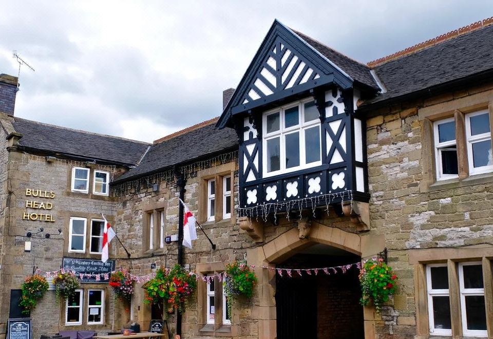 the exterior of a stone building with black and white triangular decorations , flowers , and a flag hanging from the eaves at Bulls Head