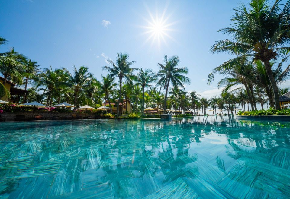 a large outdoor swimming pool surrounded by palm trees , with the sun shining brightly in the background at Pandanus Resort