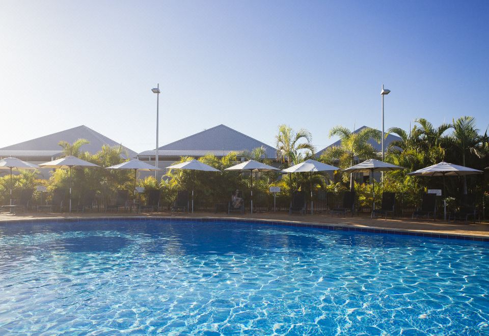 a large swimming pool with blue water and white umbrellas , surrounded by palm trees and buildings at Exmouth Escape Resort