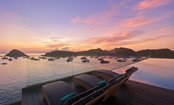 a wooden deck with two lounge chairs overlooking a body of water , surrounded by mountains and boats in the distance at Meruorah Komodo Labuan Bajo