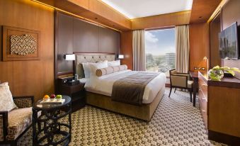 a large bed with a wooden headboard is in the center of a room with a window and a chair at Ayla Grand Hotel Al Ain