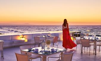 a woman in a red dress standing on a rooftop patio with a beautiful ocean view , enjoying the sunset at Grecotel Lux.ME White Palace