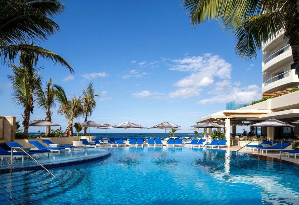 a large , blue swimming pool surrounded by palm trees and lounge chairs , with a view of the ocean in the background at Condado Vanderbilt Hotel