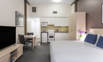 a hotel room with a bed , desk , and kitchenette area , all decorated in white and gray colors at Oaks Sydney Goldsbrough Suites
