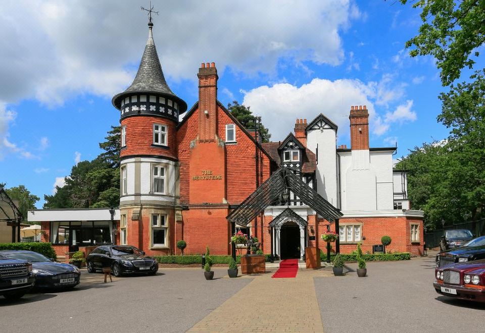 a large , red brick building with a white chimney and black roof , surrounded by trees and cars at Macdonald Berystede Hotel & Spa