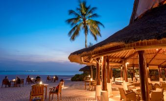 a beachside restaurant with people sitting at tables and chairs , under a thatched roof , under a blue sky with palm trees at Banyan Tree Vabbinfaru