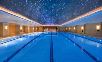The hotel features a spacious indoor swimming pool with blue and white tile walls, floor-to-ceiling windows offering a panoramic view of the city skyline, and inviting lounge chairs for guests to unwind at Radisson Blu Forest Manor Shanghai Hongqiao