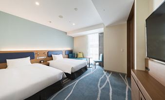 a hotel room with two beds , a desk , and a window , all set against blue walls at JR-EAST HOTEL METS FUNABASHI