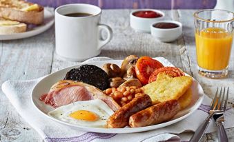 a plate of breakfast food , including bacon , eggs , sausage , mushrooms , and potatoes , is placed on a wooden table next to a cup at Premier Inn Burton On Trent East