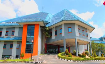 a modern building with an orange and gray exterior , topped by a blue roof , is surrounded by greenery and has a sign on the entrance at Grand Elty Singgasana Hotel