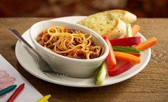 a white bowl filled with spaghetti , topped with meat sauce , sits on a dining table with a plate of bread and vegetables nearby at Premier Inn Burton On Trent East