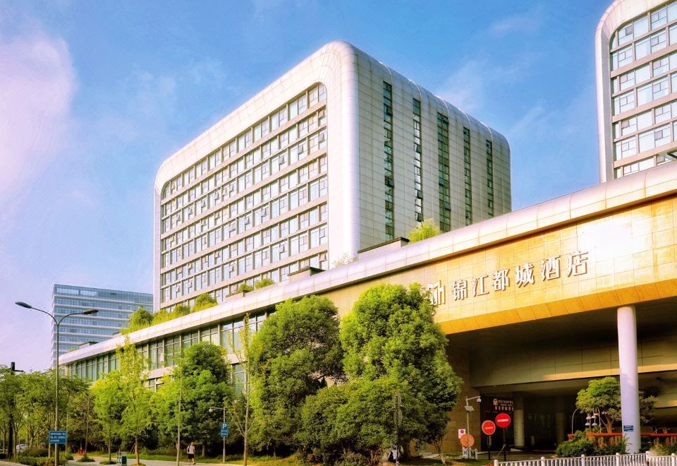 There is a front view of a street with an office building in the background, a large white building on the left side, a few parked cars in front, and a clear blue sky above at Metropolo Jinjiang Hotels (Hangzhou East Railway Station)