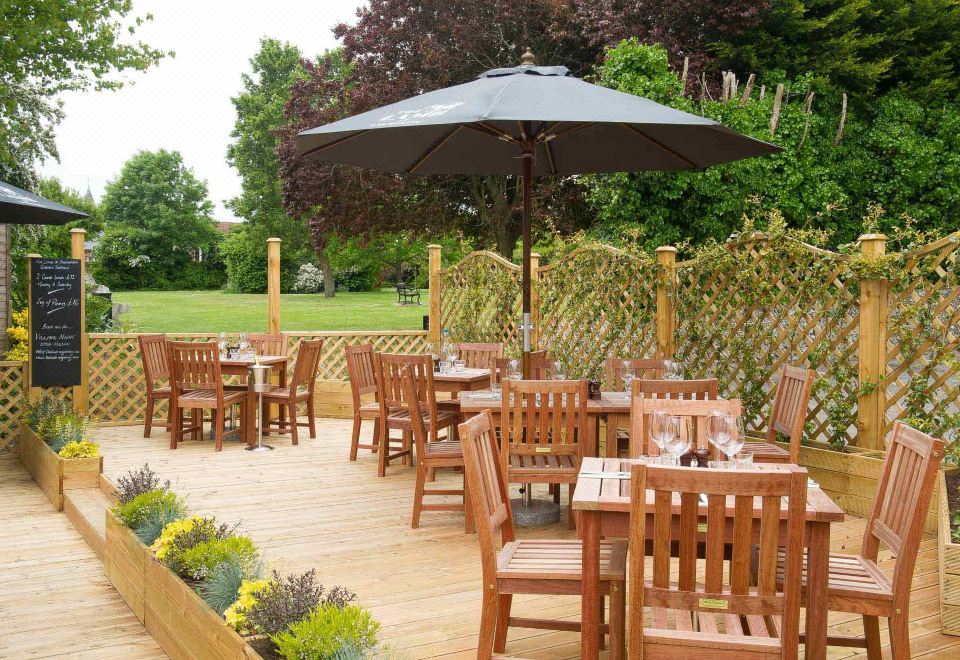 a wooden patio with tables and chairs , an umbrella , and a garden in the background at The Lamb at Angmering