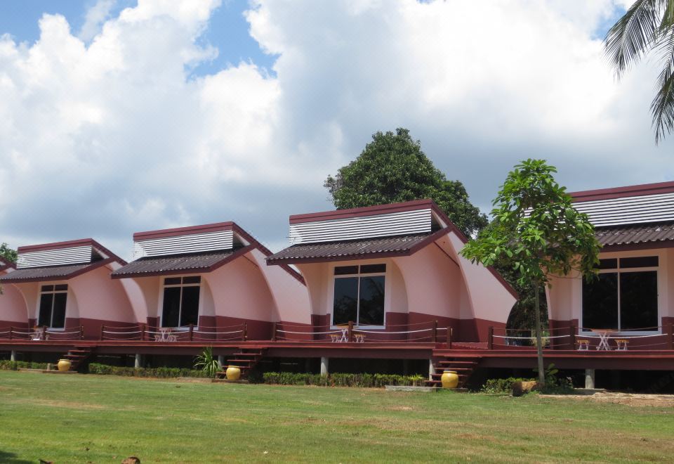 a group of pink and red wooden houses , each with a balcony , situated on a grassy field under a cloudy sky at Lomtalay Resort Trat