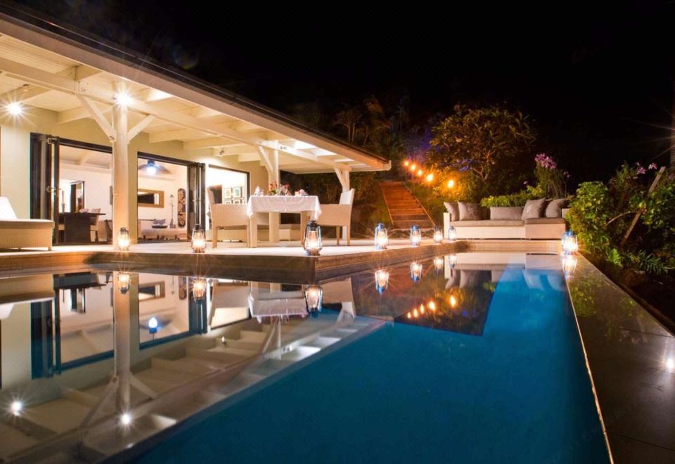 a well - lit swimming pool area at night , with a bar and lounge chairs visible in the foreground at Taveuni Palms Resort