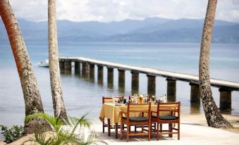 a dining table set up on a wooden deck , overlooking a body of water with mountains in the background at Jean-Michel Cousteau Resort Fiji