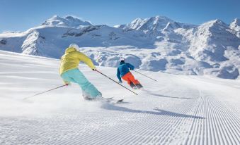 two people are skiing down a snow - covered slope , with one of them wearing a colorful outfit at Hotel Ambassador Zermatt