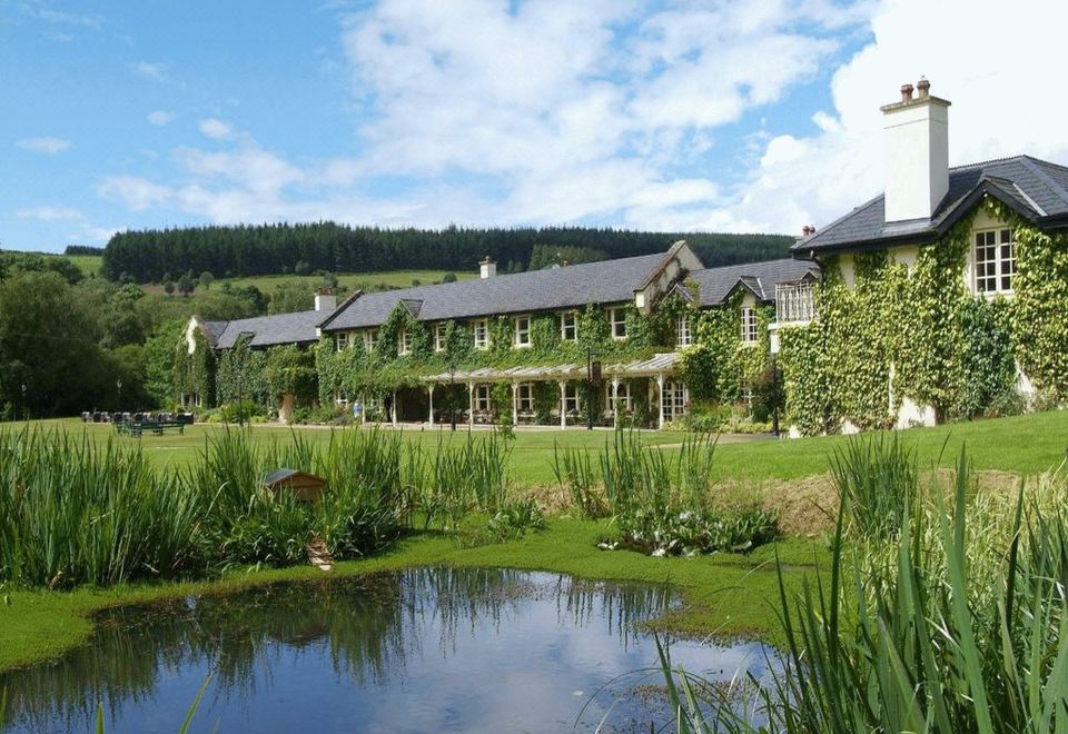 a large building with a stone facade and ivy - covered roof is situated next to a pond at BrookLodge & Macreddin Village