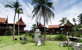 a tropical resort with wooden buildings , palm trees , and a grassy area under a cloudy sky at Maikaew Damnoen Resort