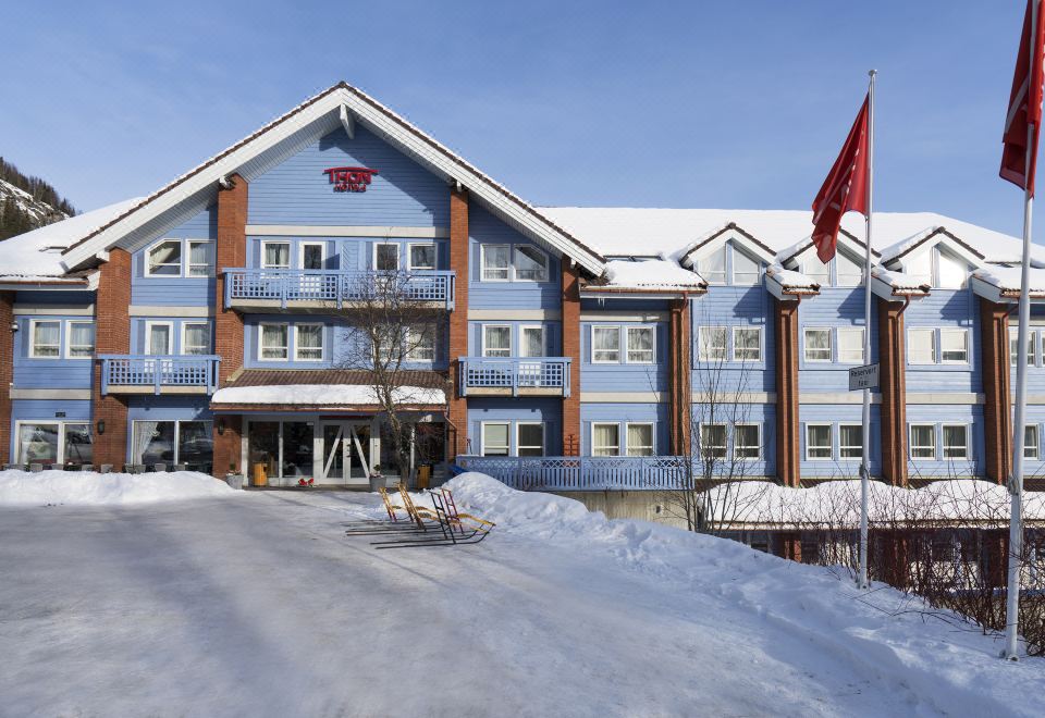 a large blue building with a red flag on top is surrounded by snow and has a clear sky at Thon Hotel Hallingdal