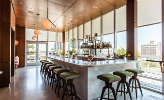 a modern bar with a long wooden counter , multiple stools , and a variety of bottles and glasses at The Dewberry Charleston
