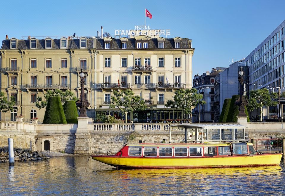 a large building with a red flag on top is reflected in the water , while a boat travels by at Hotel d'Angleterre