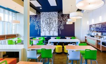 a modern , colorful restaurant with wooden tables and chairs , a mural on the wall , and pendant lights hanging from the ceiling at Ibis Budget Auckland Airport