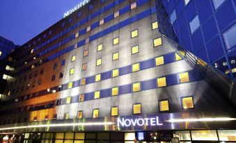 a large , modern hotel building with multiple floors , surrounded by trees and other buildings in the evening at Novotel Marne la Vallee Noisy le Grand Hotel