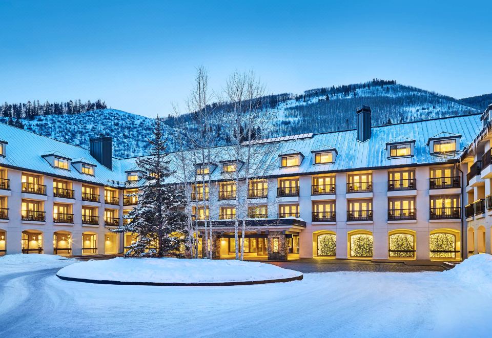 a large , well - lit hotel building with multiple stories and balconies , surrounded by snow - covered mountains at dusk at Grand Hyatt Vail