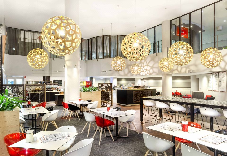 a modern restaurant with white tables and chairs , red accents , and large pendant lights hanging from the ceiling at Ibis Melbourne Hotel and Apartments