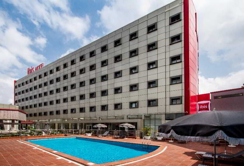 a large red hotel with a pool and lounge chairs , under a blue sky with clouds at Ibis Lagos Ikeja