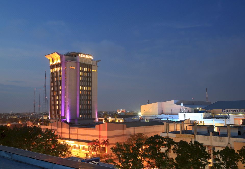 a tall building with purple lights is illuminated at night , and cars are parked nearby at Aryaduta Palembang