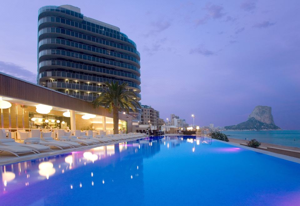 a large swimming pool surrounded by buildings and trees , with the sun setting in the background at Solymar Gran Hotel