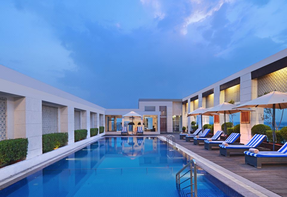 a large outdoor swimming pool surrounded by multiple buildings , with lounge chairs and umbrellas placed around the pool area at Radisson Hotel Agra