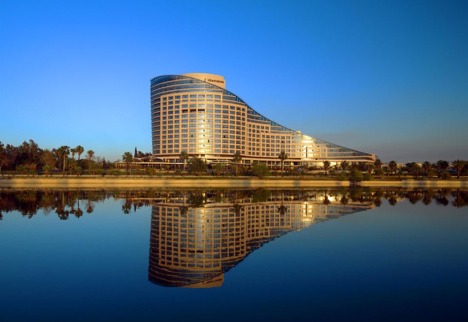 a large , modern hotel building surrounded by a body of water , with the sun setting in the background at Sheraton Grand Adana