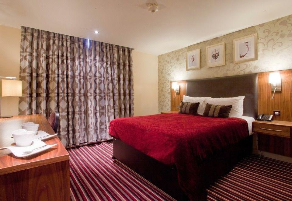 a large bed with a red blanket is in the middle of a room with a desk , chair , and curtains at Gloucester Robinswood Hotel, BW Signature Collection