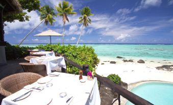 a tropical beachfront restaurant with white tables and chairs , palm trees , and a clear blue sky at Small Luxury Hotels of the World - Pacific Resort Aitutaki