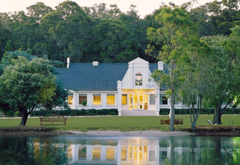 a large white house surrounded by trees , with a pond in the foreground and a lake in the background at Cape Lodge