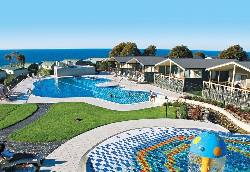 a large outdoor swimming pool surrounded by grass and trees , with people enjoying themselves in the water at NRMA Merimbula Beach Holiday Resort