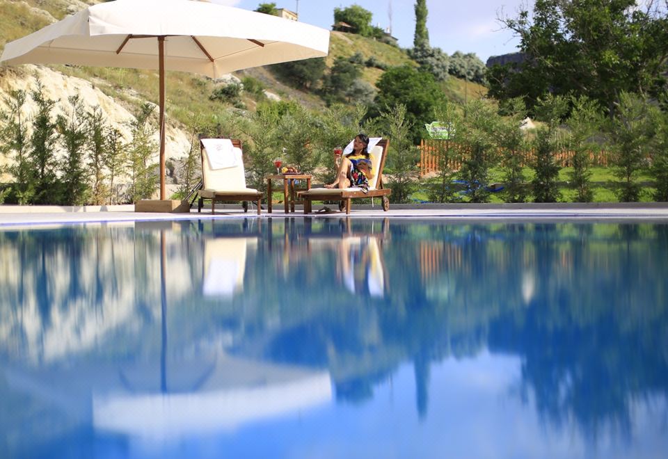 a woman sitting on a lounge chair by a large blue pool , surrounded by lush greenery at Kapadokya Hill Hotel & Spa (12+)