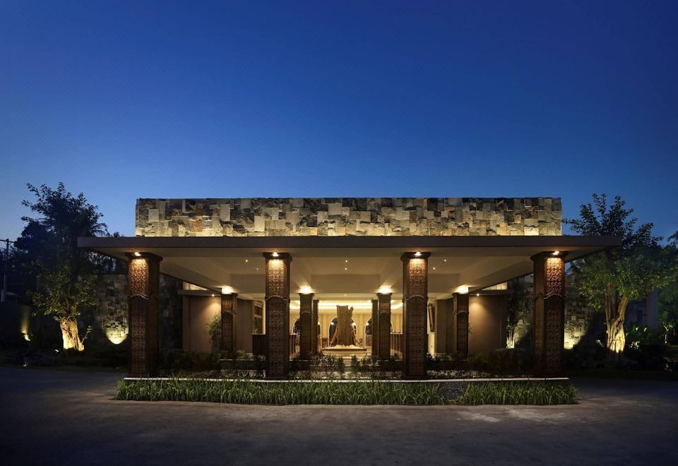 a large building with a stone facade and columns is lit up at night , surrounded by greenery at The Westlake Hotel & Resort Yogyakarta
