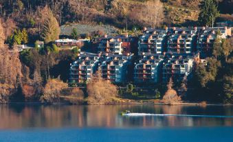 a row of multi - story buildings are situated on the shore of a lake , with a boat sailing in the water below at Oaks Queenstown Shores Resort