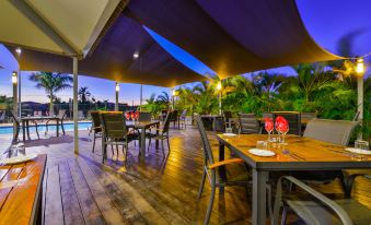 a wooden deck with tables and chairs under a large purple canopy , overlooking a pool area at dusk at Exmouth Escape Resort