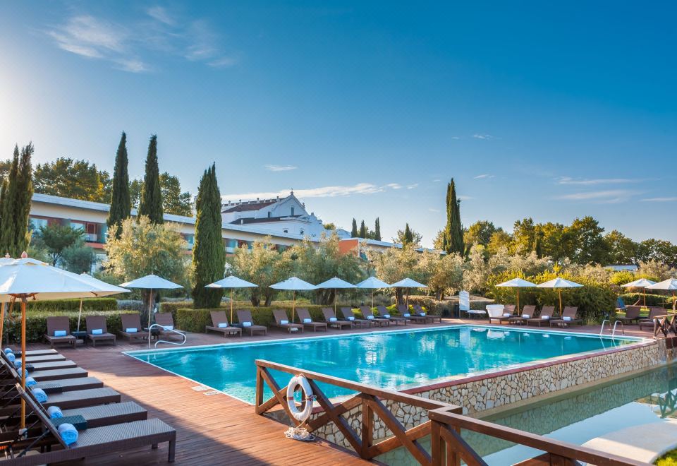 a large outdoor pool surrounded by lounge chairs and umbrellas , with a hotel in the background at Convento do Espinheiro, Historic Hotel & Spa