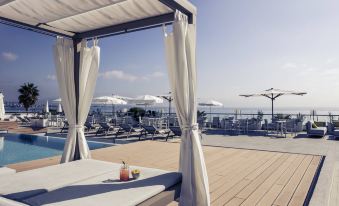 a white canopy bed on a wooden deck overlooking a pool and the ocean , with chairs and umbrellas nearby at Mercure Villeneuve Loubet Plage