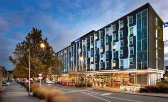a modern , glass - fronted building with multiple floors and balconies , situated on a busy street at dusk at Ibis Budget Auckland Airport