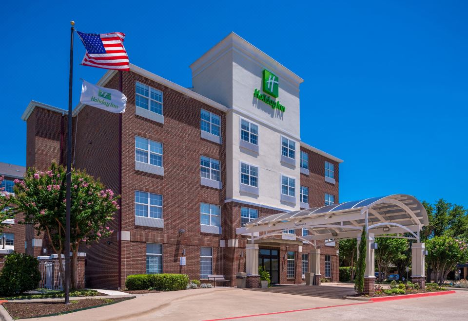 a large hotel building with a green sign and american flag in front of it at Holiday Inn & Suites Dallas-Addison